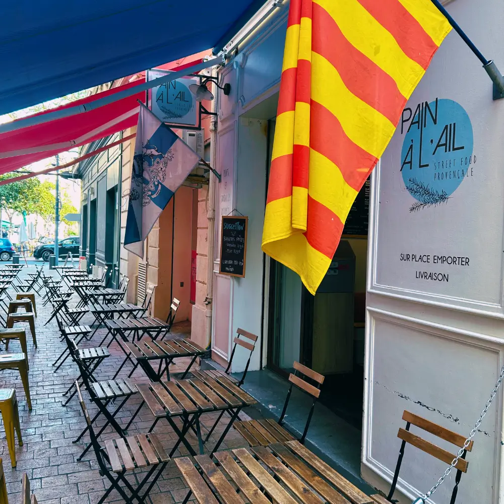Terrasse du restaurant Pain à l’Ail avec parasols colorés, table dressée et enseigne visible en façade.