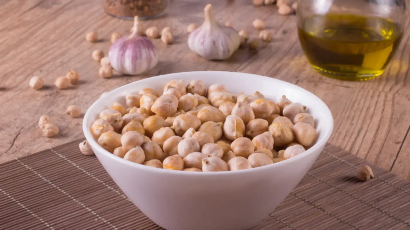 Bol de pois chiches sur une table en bois, avec ail et huile d’olive en arrière-plan.